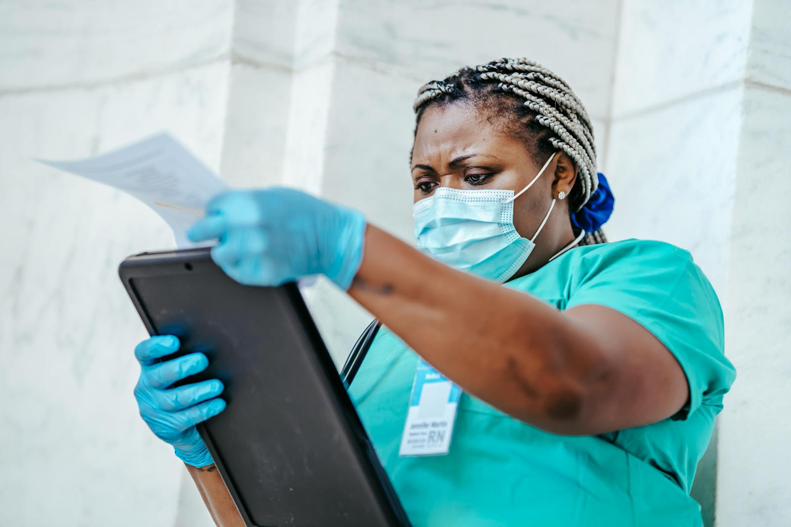 Low angle of focused black female nurse in mask and gloves standing in hospital and looking through papers