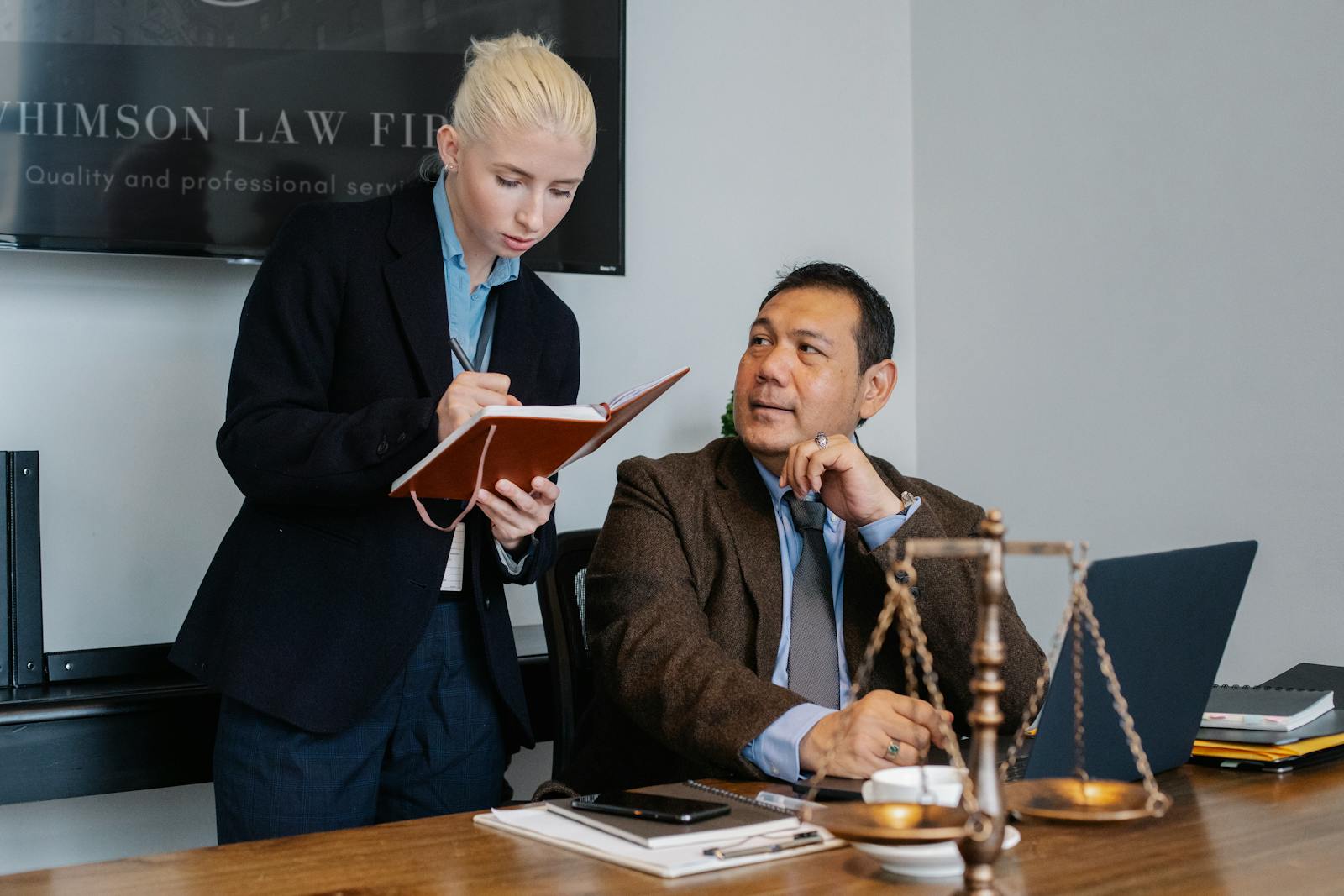 Young female secretary in formal clothes taking notes in notebook and taking to serious Asian male manager while working in law firm together